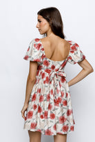 Back view of a woman wearing an ivory  dress with red rose pattern and a self-tie bow a white background