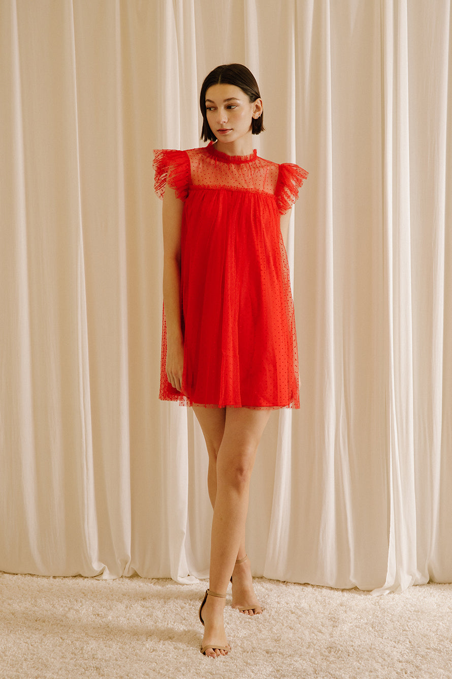 Alternative view of a woman wearing a red dress with tulle fabric with mini polka dot design throughout against a beige wall.