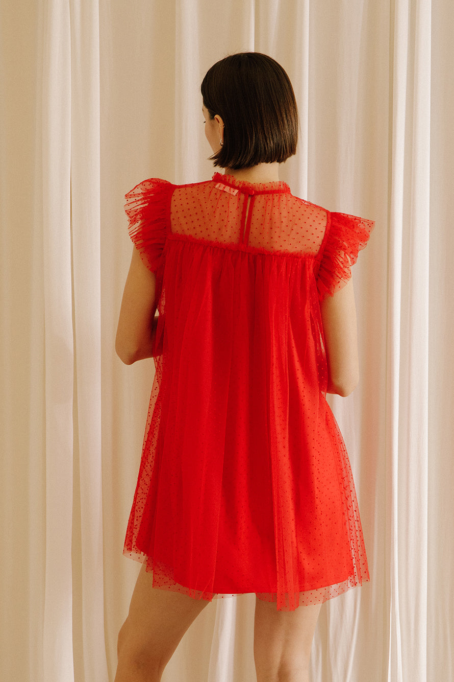 Back view of a woman wearing a red dress with tulle fabric with mini polka dot design throughout against a beige wall.