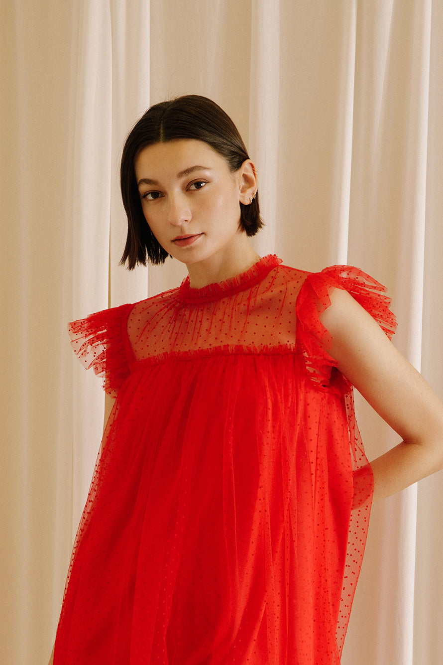 Close-up of a woman wearing a red dress with tulle fabric with mini polka dot design throughout against a beige wall.