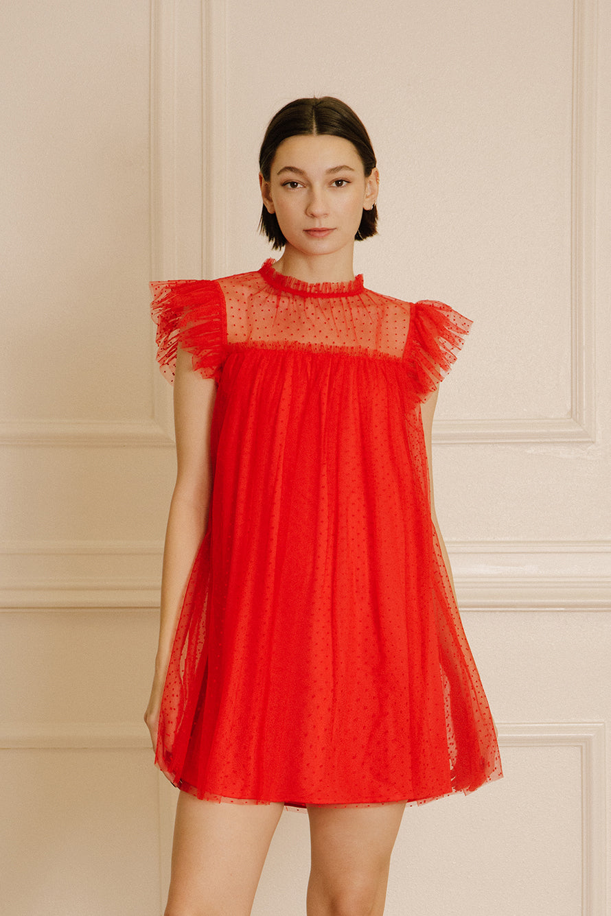 Woman wearing a red dress with tulle fabric with mini polka dot design throughout against a beige wall.