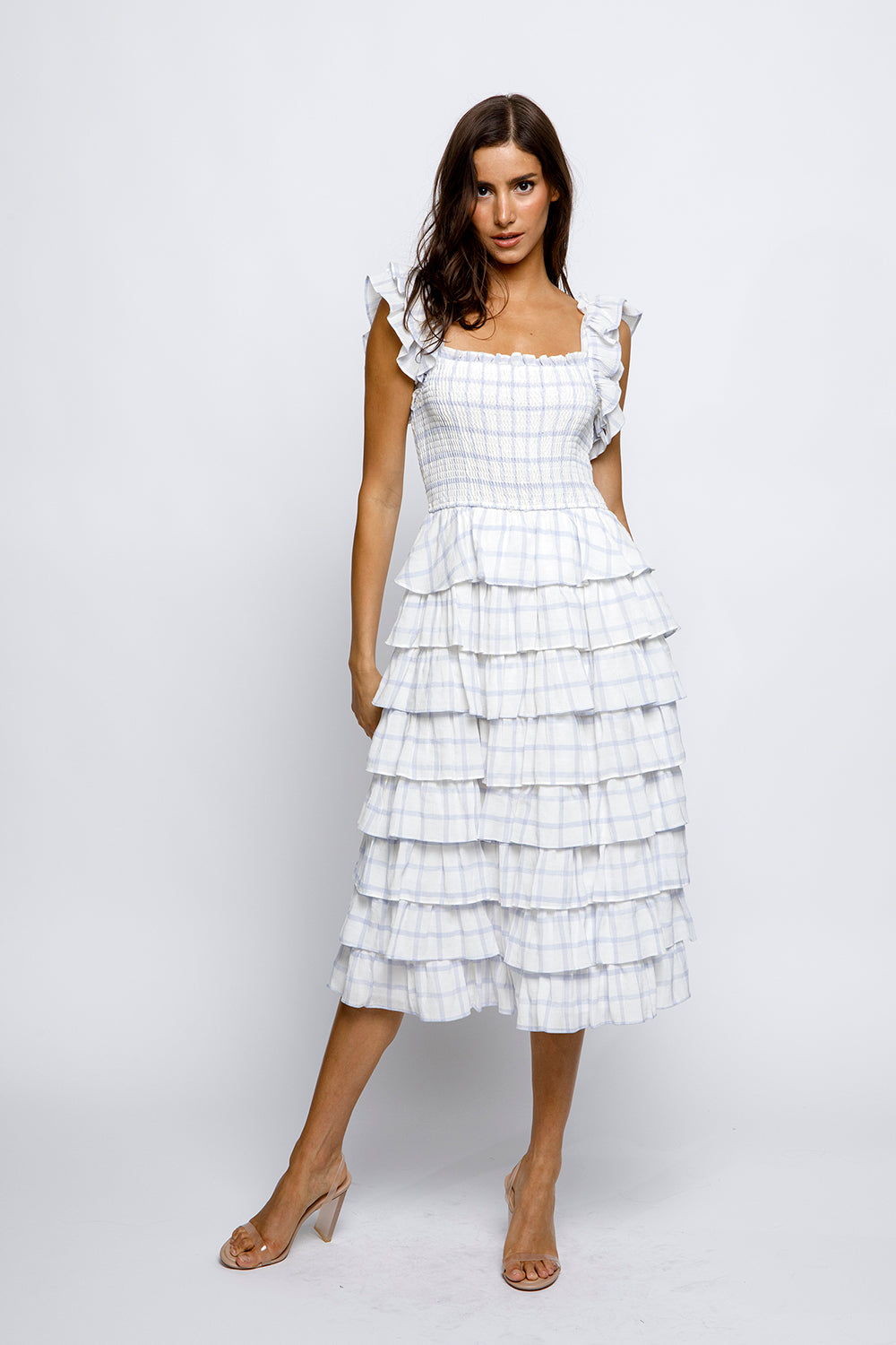 A woman wearing a white and blue gingham midi dress with a square neckline, smocked fitted upper body, and tiered ruffles. She is also wearing beige heels.