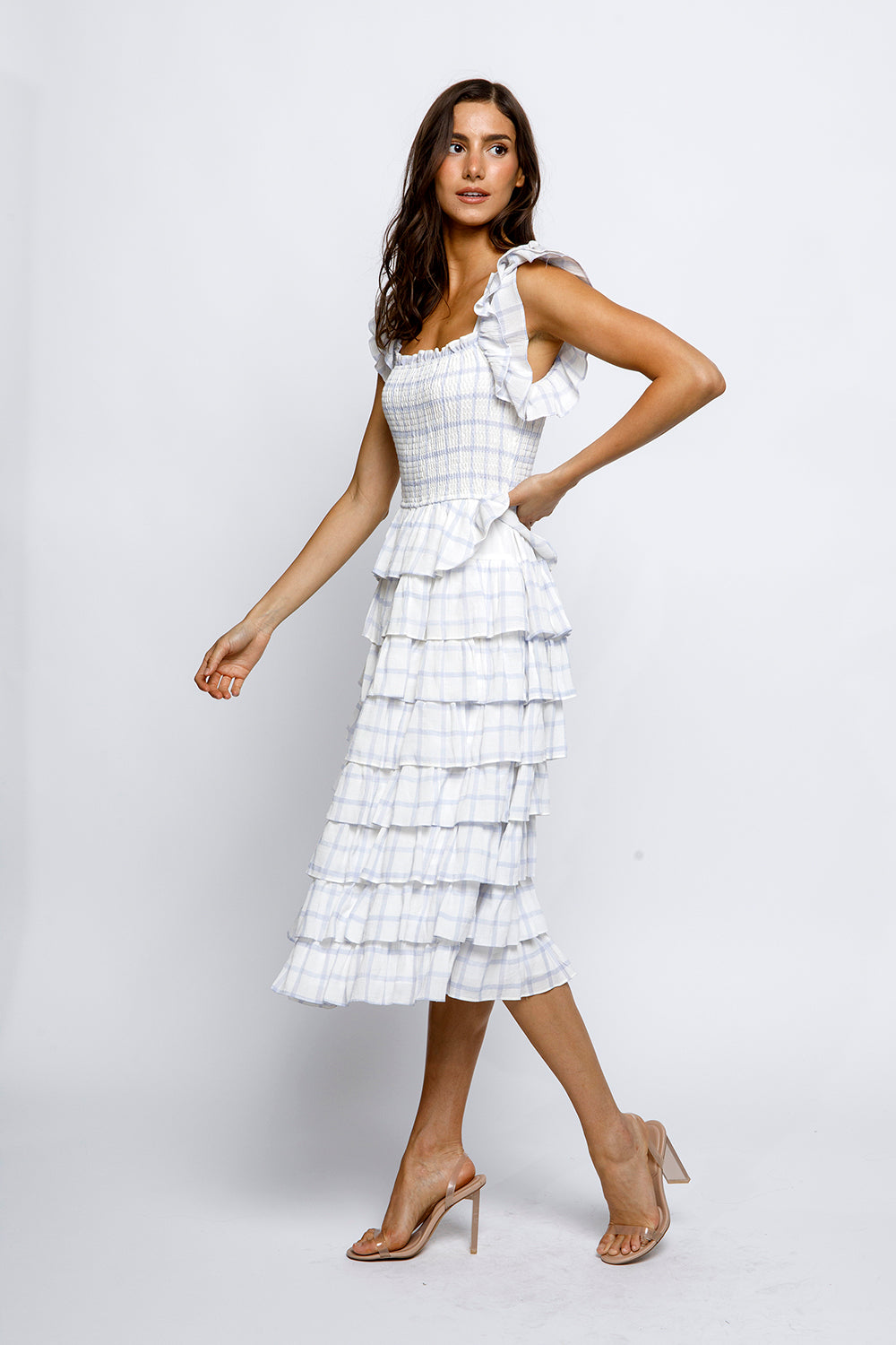 A woman wearing a white and blue gingham midi dress with a square neckline, smocked fitted upper body, ruffled capped sleeves, and tiered ruffles. She is also wearing beige heels.