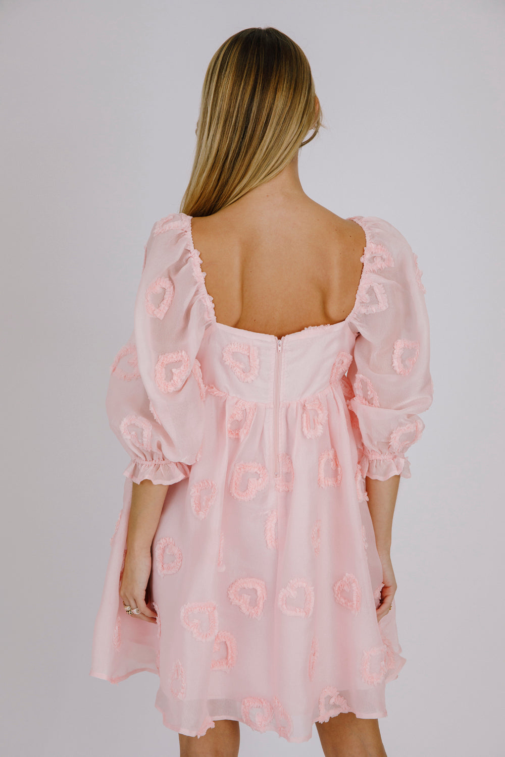 Back view of a woman wearing a pink dress with heart patterns on a plain background