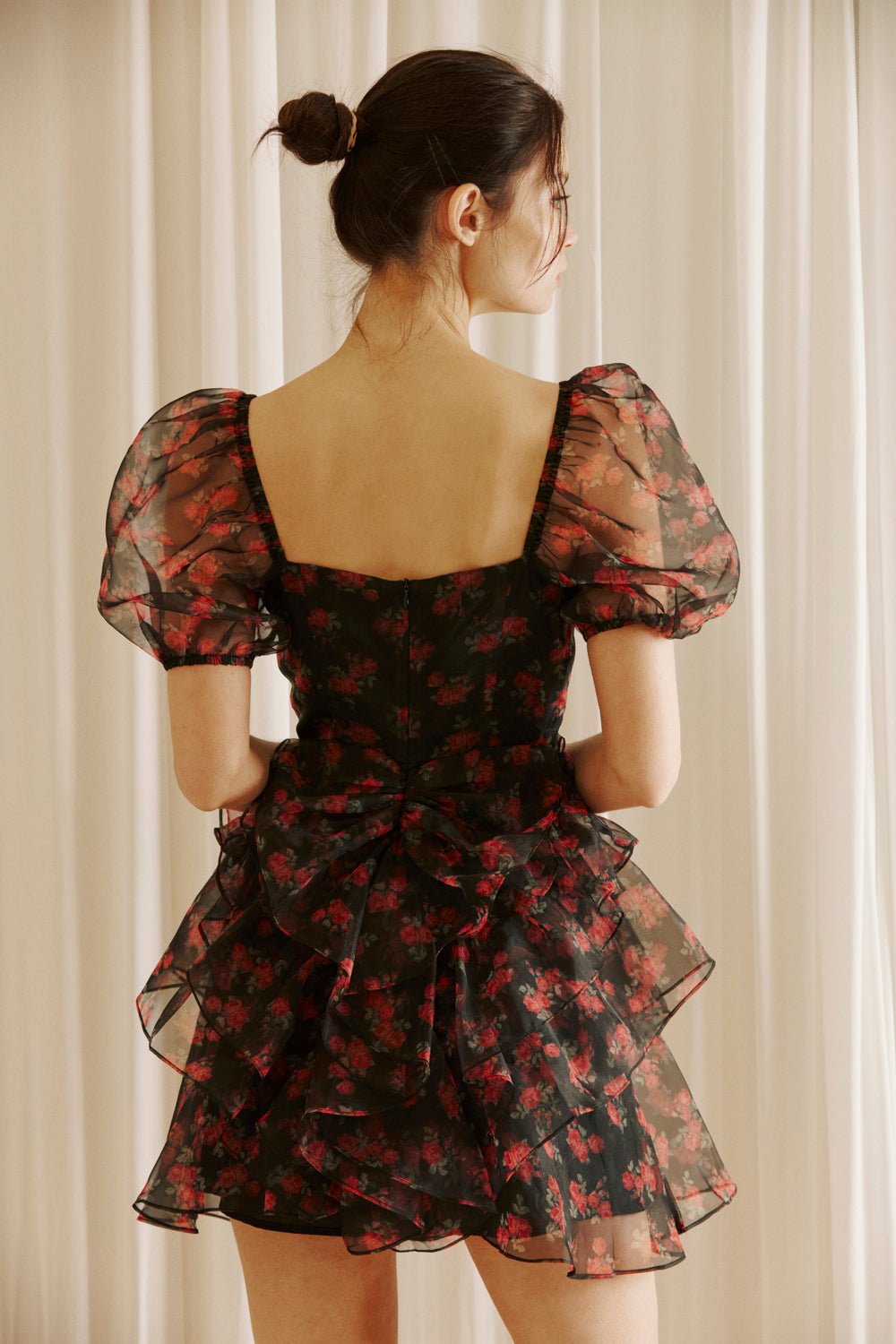 Back view of a woman wearing a black floral dress with sheer puff sleeves against a light curtain background