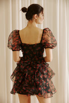 Back view of a woman wearing a black floral dress with sheer puff sleeves against a light curtain background