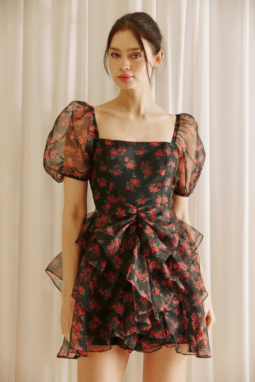 Woman wearing a black floral dress with sheer puff sleeves against a light curtain background
