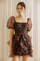 Woman wearing a black floral dress with sheer puff sleeves against a light curtain background