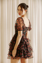Side view of a woman wearing a black floral dress with sheer puff sleeves against a light curtain background