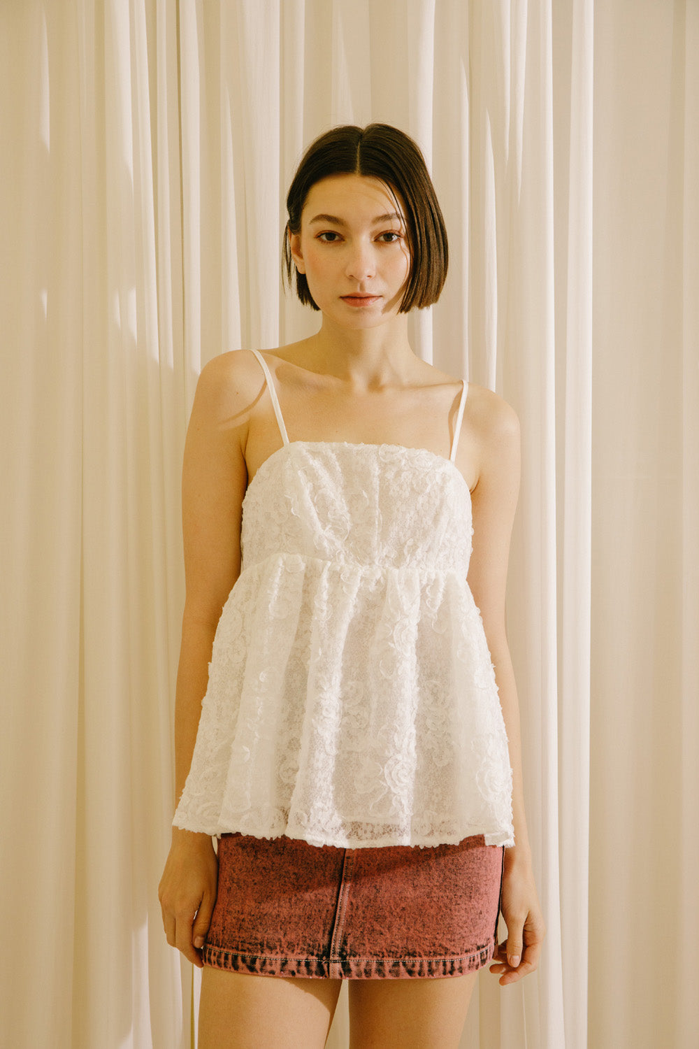 A female model wearing a white 3D floral lace top with adjustable spaghetti straps and a gathered empire waist, paired with a red skirt.