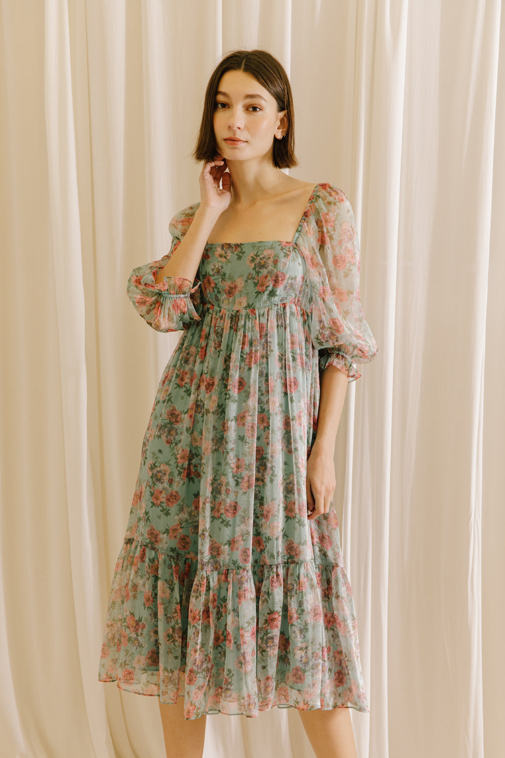 Woman wearing a teal floral midi dress against a beige curtain background