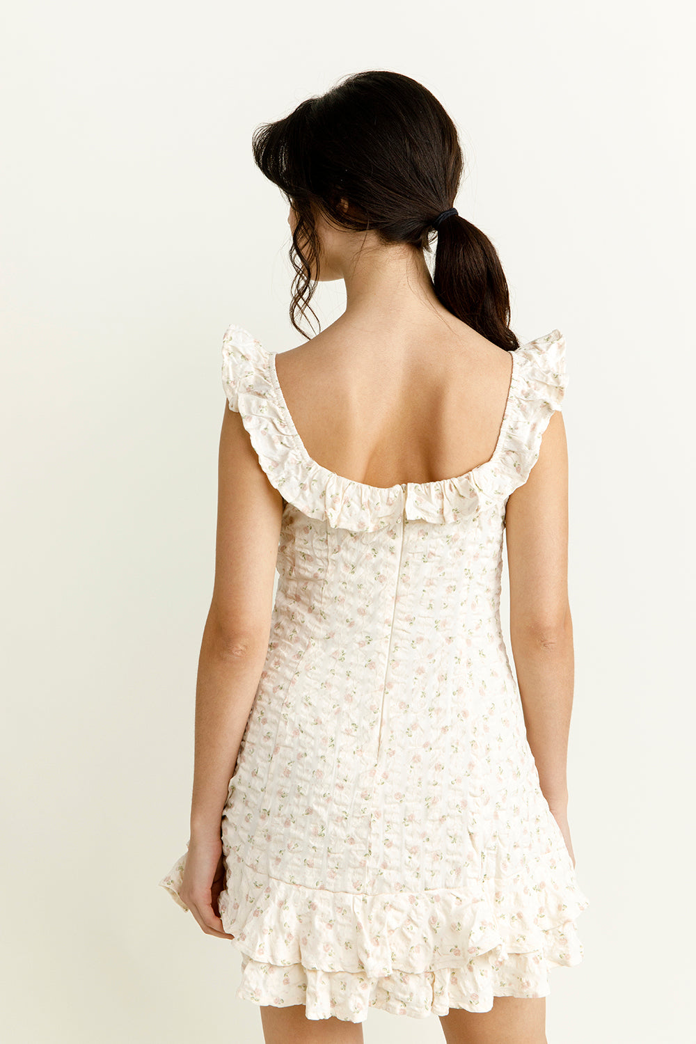 Back view of a woman wearing an ivory floral mini dress with ruffled straps against a plain background