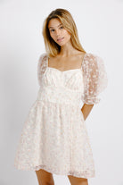 Woman wearing a cream dress with floral pattern and sheer puffy sleeves on a plain background