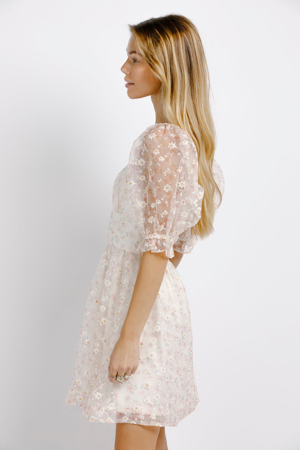 Side view of woman wearing a cream dress with floral pattern and sheer puffy sleeves on a plain background
