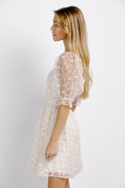 Side view of woman wearing a cream dress with floral pattern and sheer puffy sleeves on a plain background