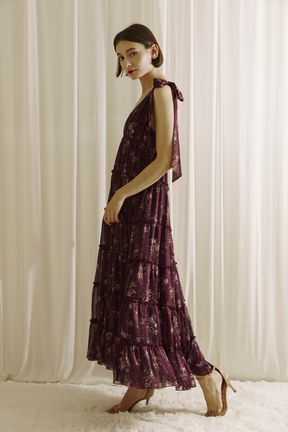 Side view of a woman modeling a plum base maxi dress with an allover pink floral print and beige heels on a white background