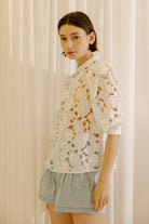 A woman wearing a white eyelet lace blouse with puff sleeves and light blue denim shorts, standing in front of a soft beige curtain backdrop.