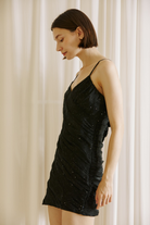 A woman wearing a black spaghetti-strap mini dress with ruching and subtle sequin embellishments, standing in front of white curtains.