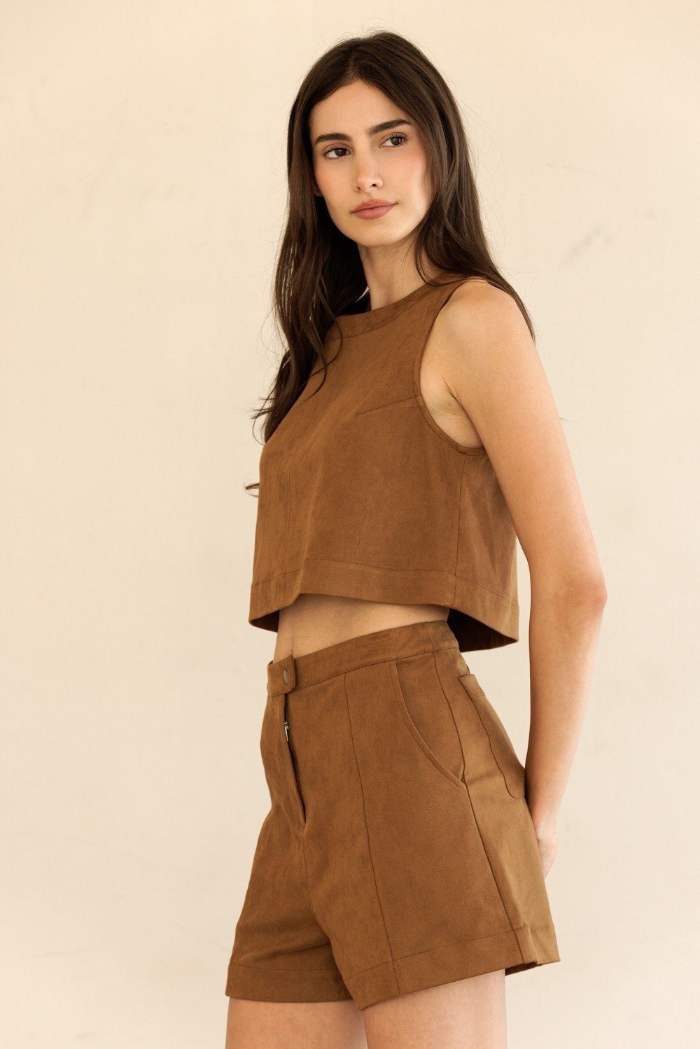 A woman modeling a matching brown faux-suede two-piece set consisting of a sleeveless crop top and high-waisted shorts, set against a plain beige background.
