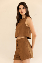 A woman modeling a matching brown faux-suede two-piece set consisting of a sleeveless crop top and high-waisted shorts, set against a plain beige background.