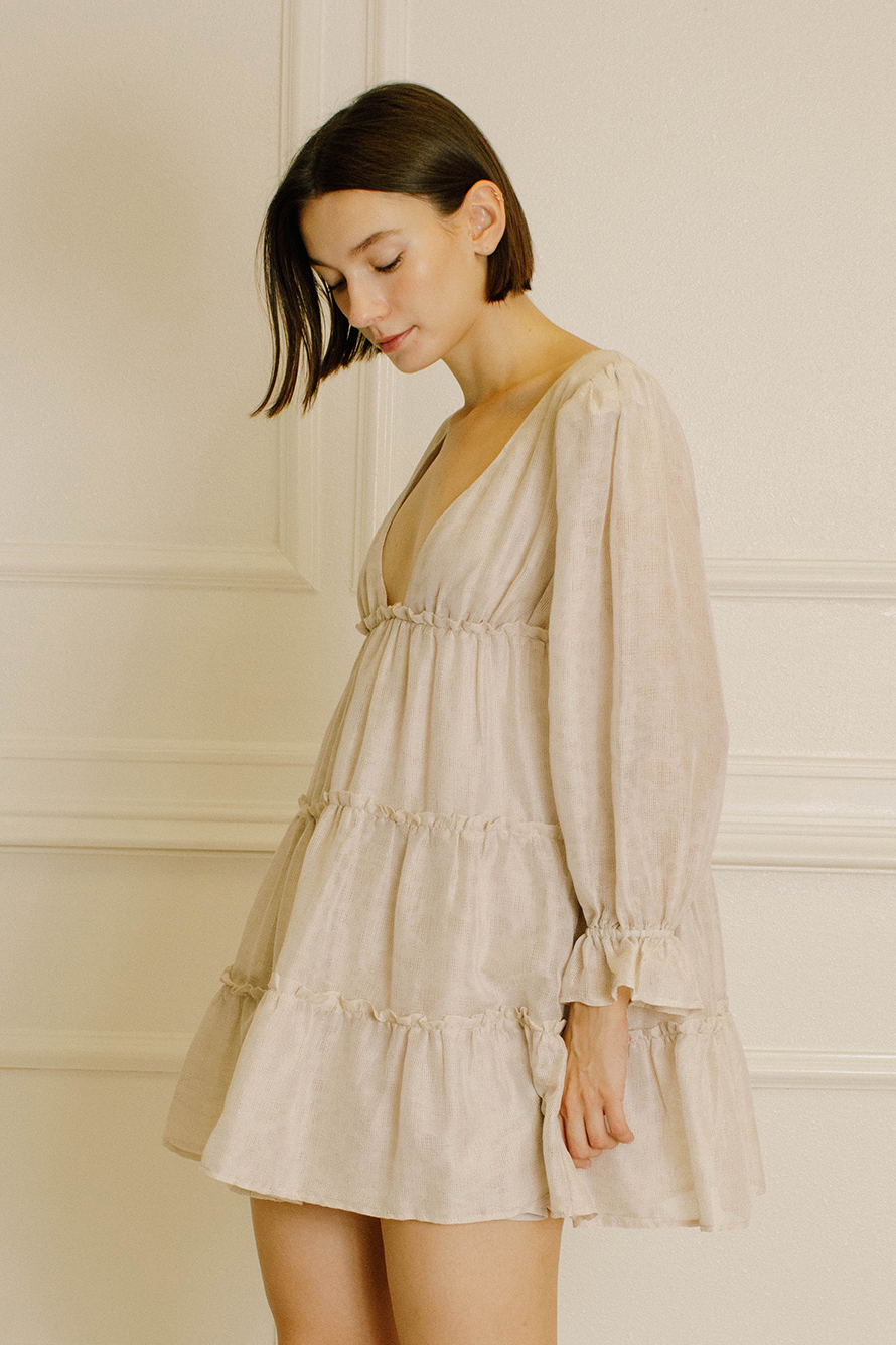 A woman wearing a light beige tiered dress with long ruffled sleeves, standing against a cream-colored paneled wall.