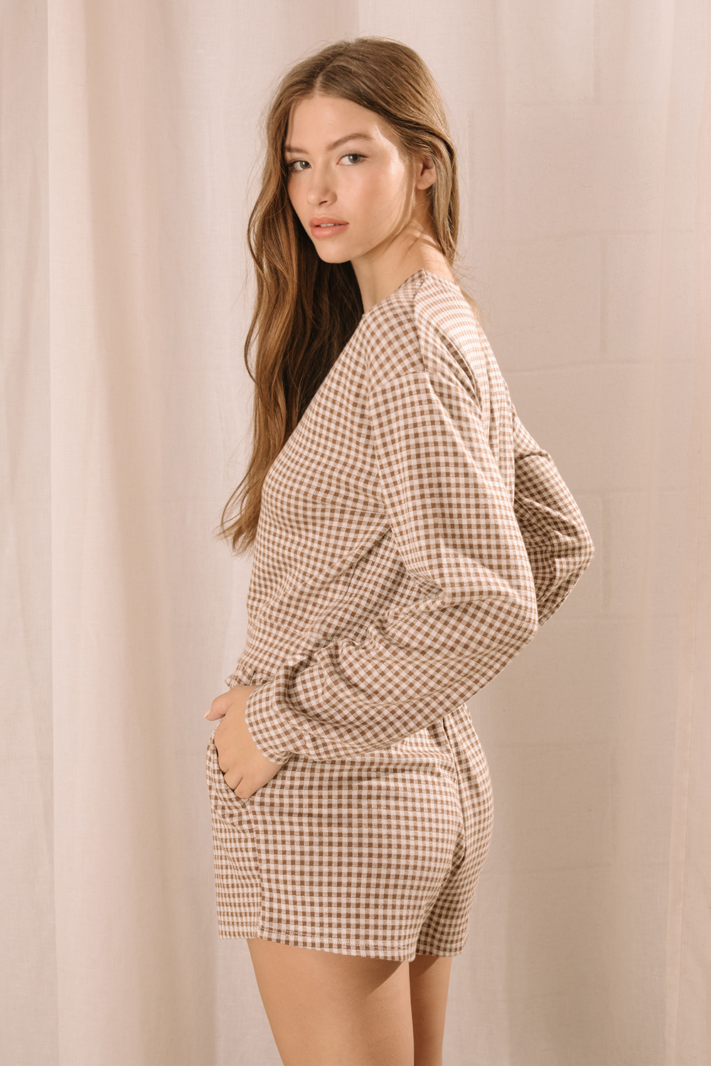 Side view of a woman modeling a lounge set that features a long-sleeve top and high-waisted drawstring shorts with pockets in a brown and ivory gingham print.