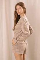Side view of a woman modeling a lounge set that features a long-sleeve top and high-waisted drawstring shorts with pockets in a brown and ivory gingham print.