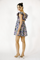 Side view of a model wearing a colorful floral mini dress featuring a ruffled cap sleeves, flowy silhouette, and open bow-tie back.