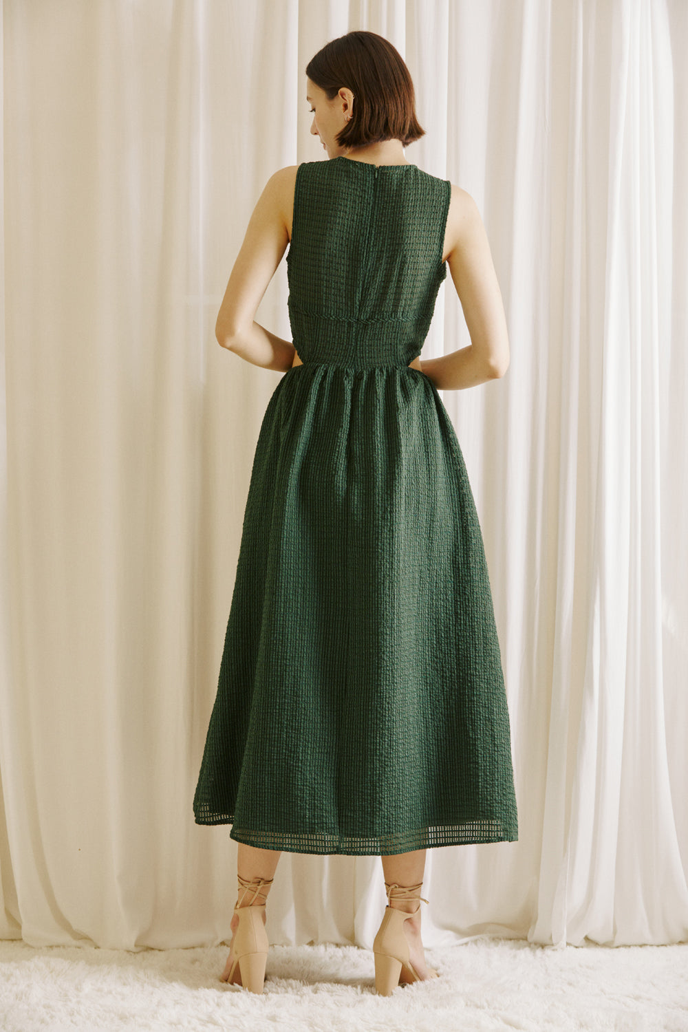 Back view of a woman wearing a green midi dress with side cut-outs and beige high-heels standing against a white curtain backdrop