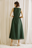 Back view of a woman wearing a green midi dress with side cut-outs and beige high-heels standing against a white curtain backdrop