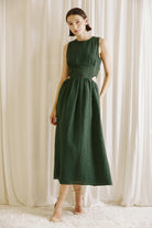 Woman wearing a green midi dress with side cut-outs and beige high-heels standing against a white curtain backdrop