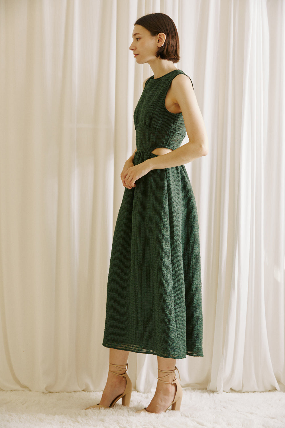 Side view of a woman wearing a green midi dress with side cut-outs and beige high-heels standing against a white curtain backdrop