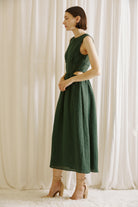Side view of a woman wearing a green midi dress with side cut-outs and beige high-heels standing against a white curtain backdrop