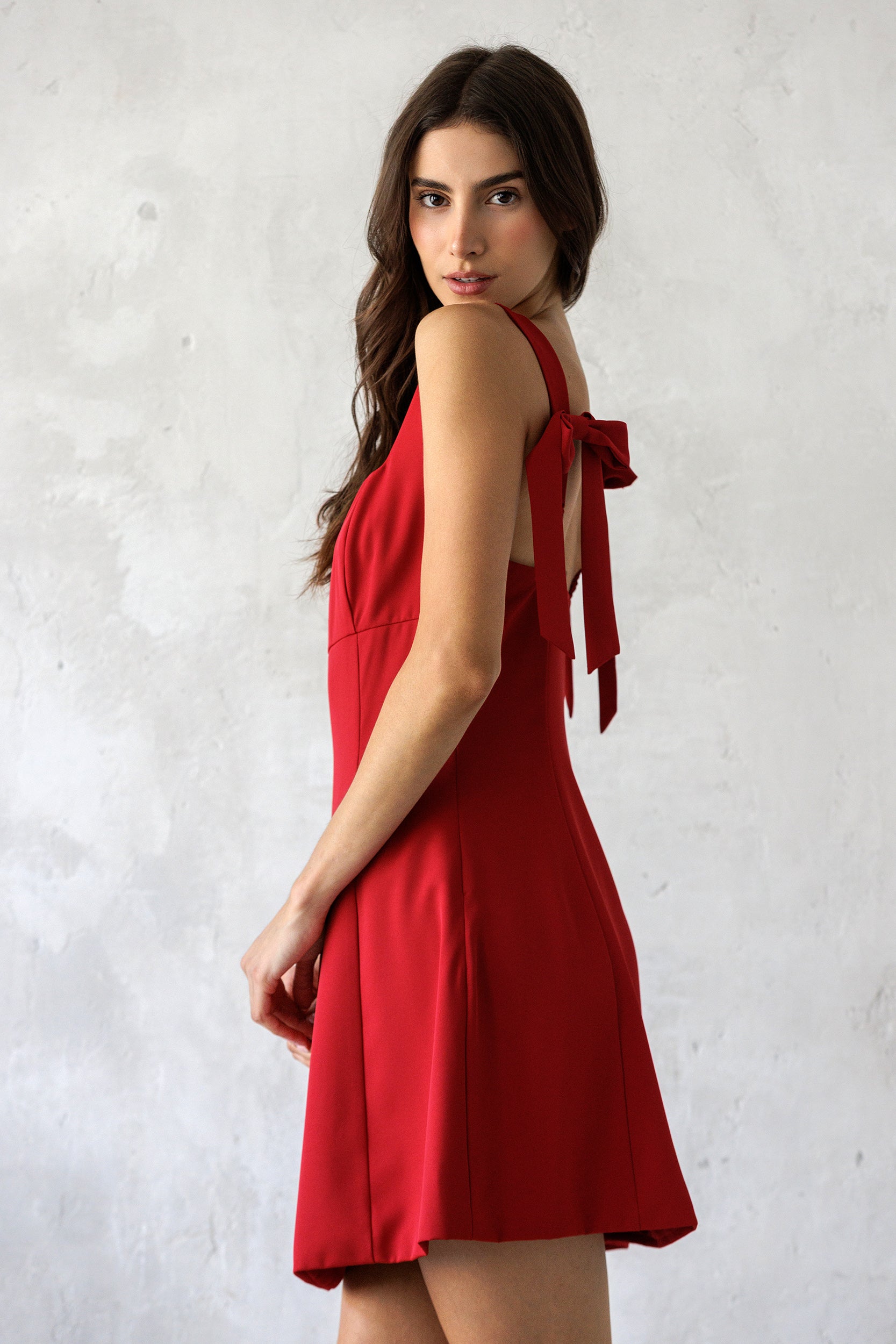A side view of a model wearing a timeless red mini dress featuring a square neckline, fit-and-flare silhouette, and tie shoulder straps for an effortlessly chic look.