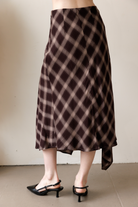 A woman viewed from behind wearing a brown and beige plaid midi skirt that features a bias-cut silhouette and a flowy asymmetrical handkerchief hem.