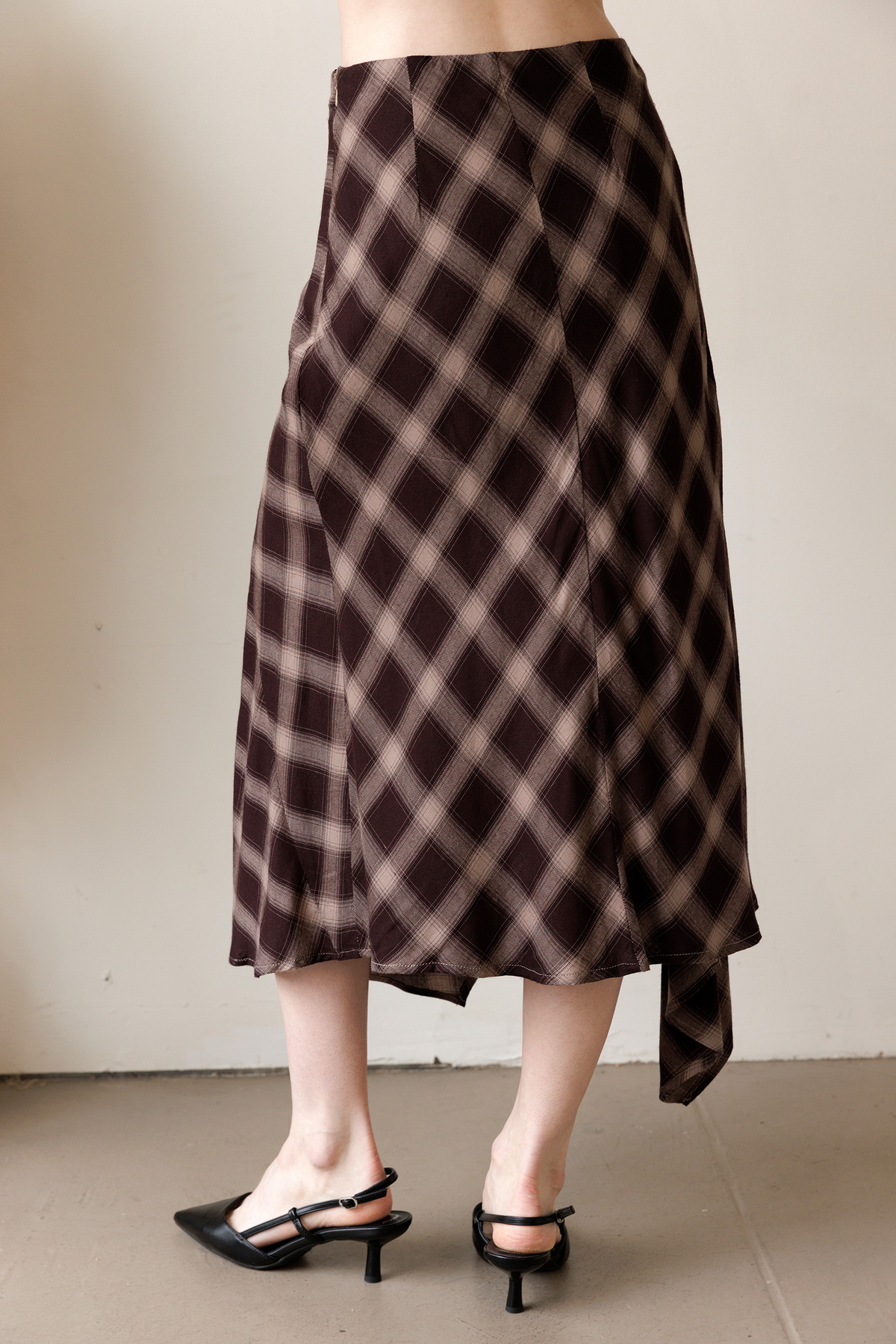 A woman viewed from behind wearing a brown and beige plaid midi skirt that features a bias-cut silhouette and a flowy asymmetrical handkerchief hem.