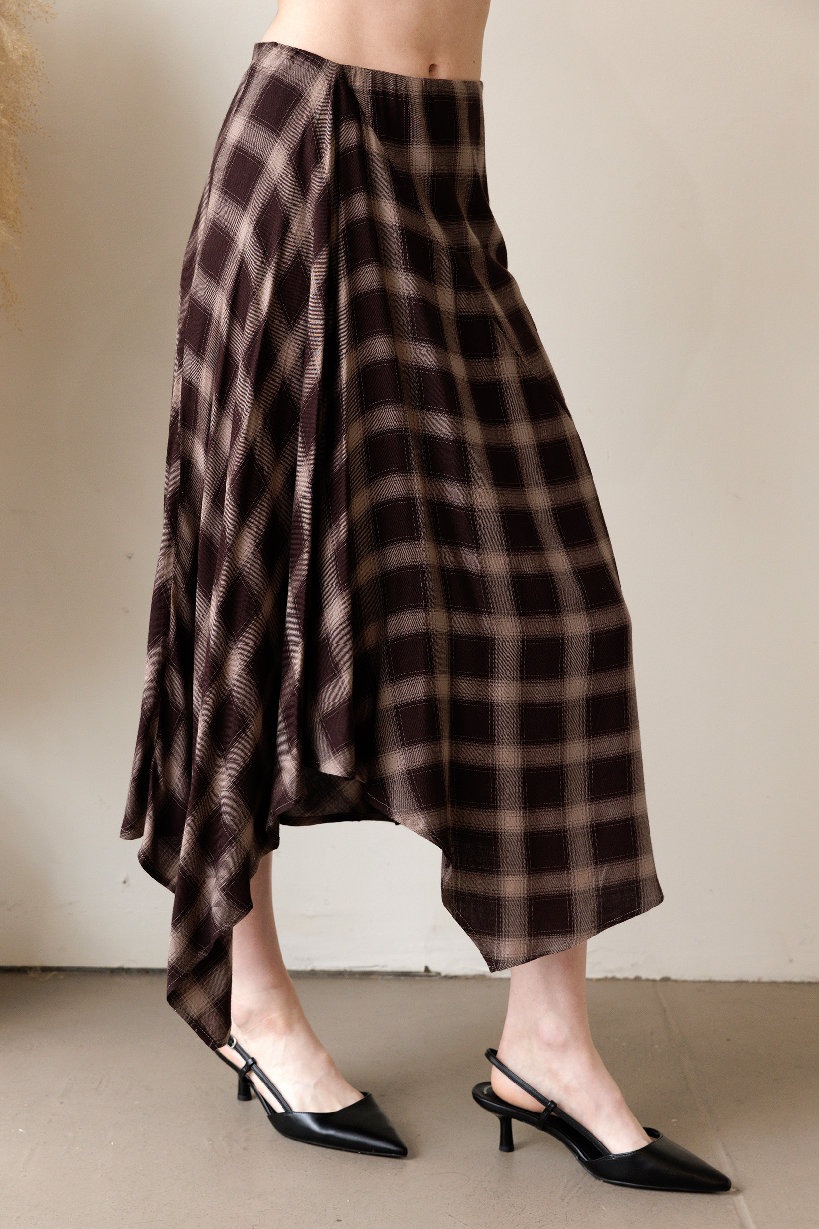 A person wearing a plaid midi skirt that features a brown and beige print and a flowy asymmetrical handkerchief hem.