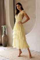 Woman wearing a yellow maxi dress standing in a room with a decorative vase and plant.