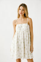 A woman standing in a studio setting, wearing a white sleeveless mini dress with blue and green floral print, spaghetti straps, and a square neckline.