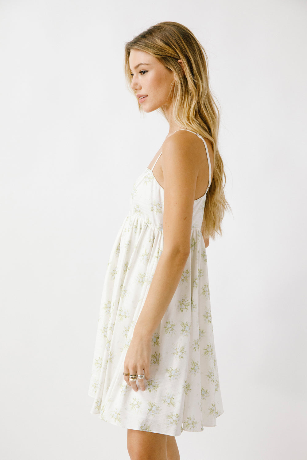 Side view of a woman standing in a studio setting, wearing a white sleeveless mini dress with blue and green floral print, spaghetti straps, and a square neckline.