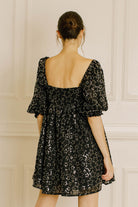 Woman wearing a black mini dress with a backless design against a white paneled wall.