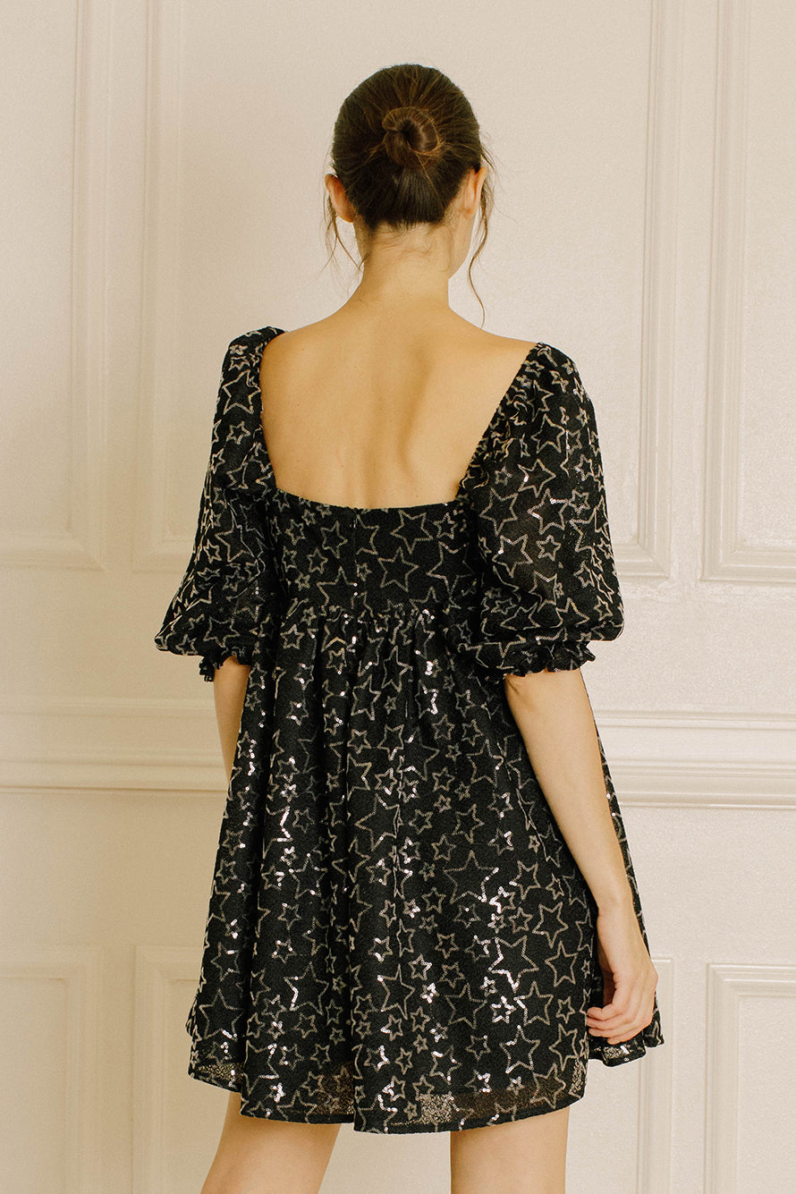 Woman wearing a black mini dress with a backless design against a white paneled wall.