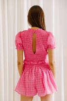 Back view of a woman wearing a pink romper against a white curtain background