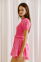 Side view of a woman wearing a pink romper against a white curtain background