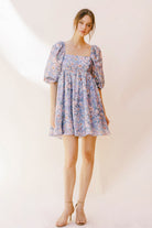 Woman wearing a purple paint stroke print dress with beige high-heels against a plain background