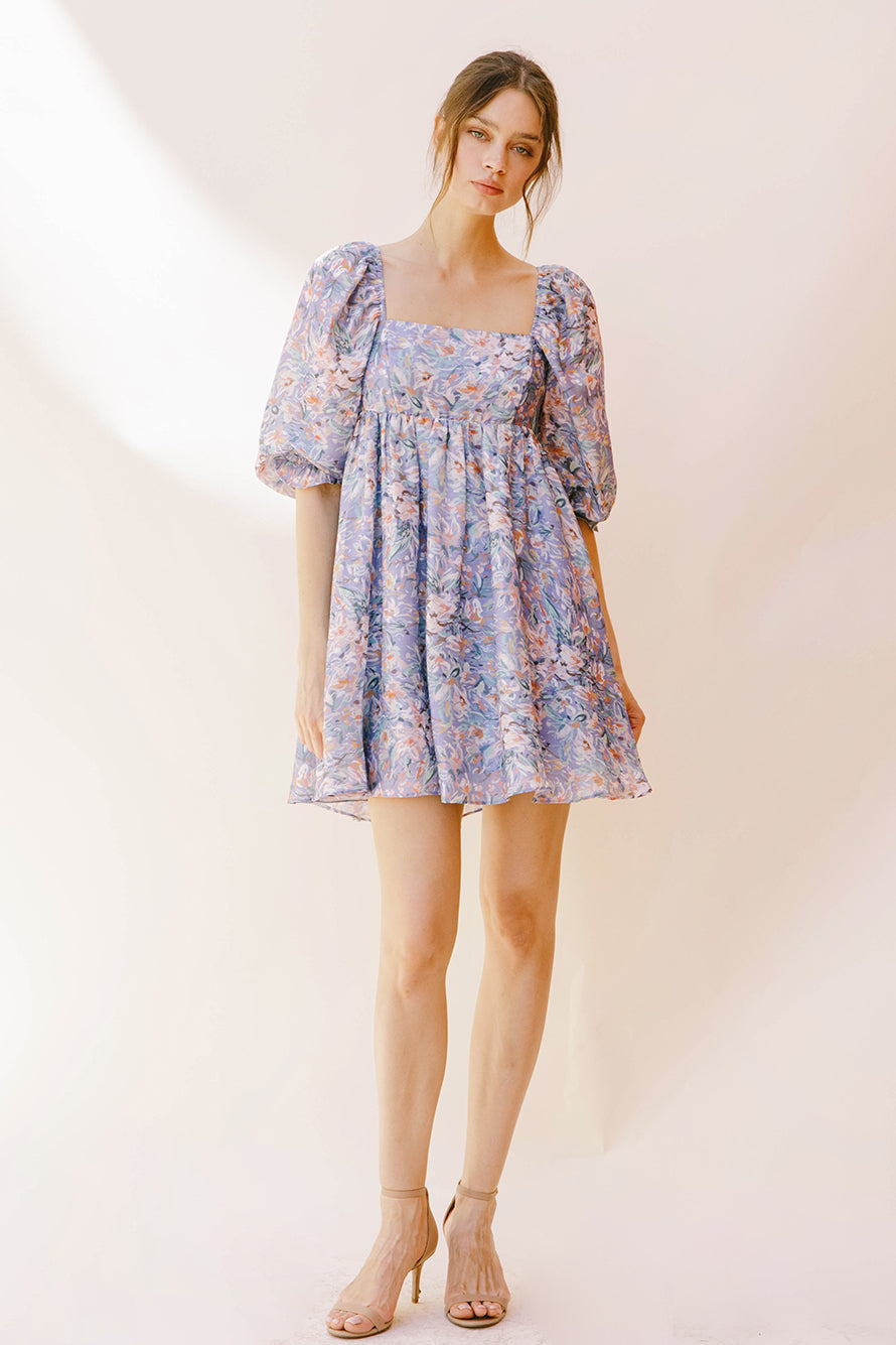 Woman wearing a purple paint stroke print dress with beige high-heels against a plain background