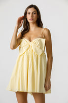 A woman wearing a yellow mini dress with an oversized front bow, spaghetti straps, and a sweetheart neckline.