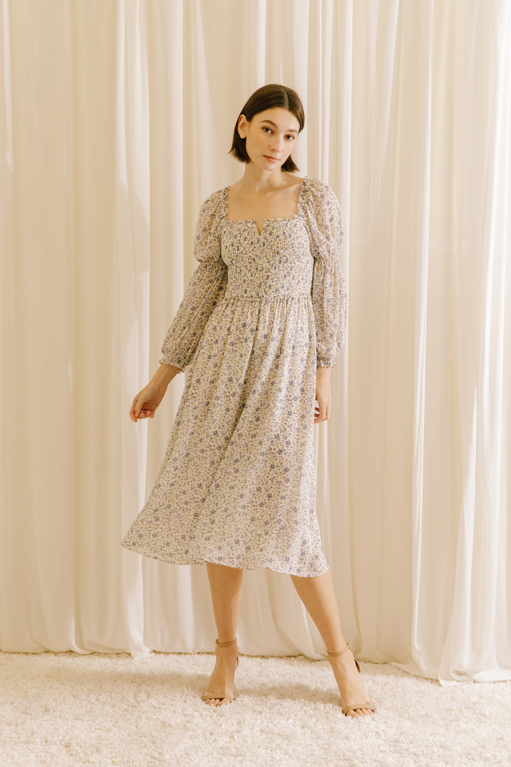 A woman standing in a room with curtains wearing a beige floral square neckline midi dress with long sleeves.
