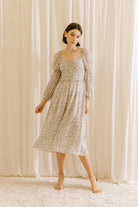A woman standing in a room with curtains wearing a beige floral square neckline midi dress with long sleeves.
