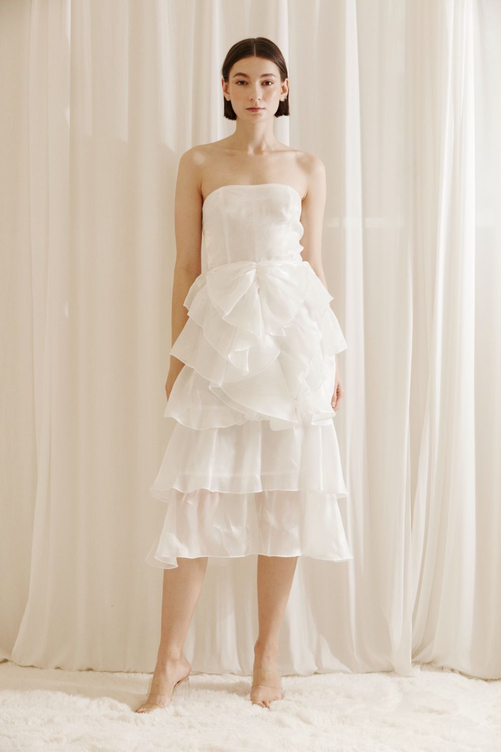 Woman wearing a white strapless dress with asymmetrical ruffled layers against a white curtain background